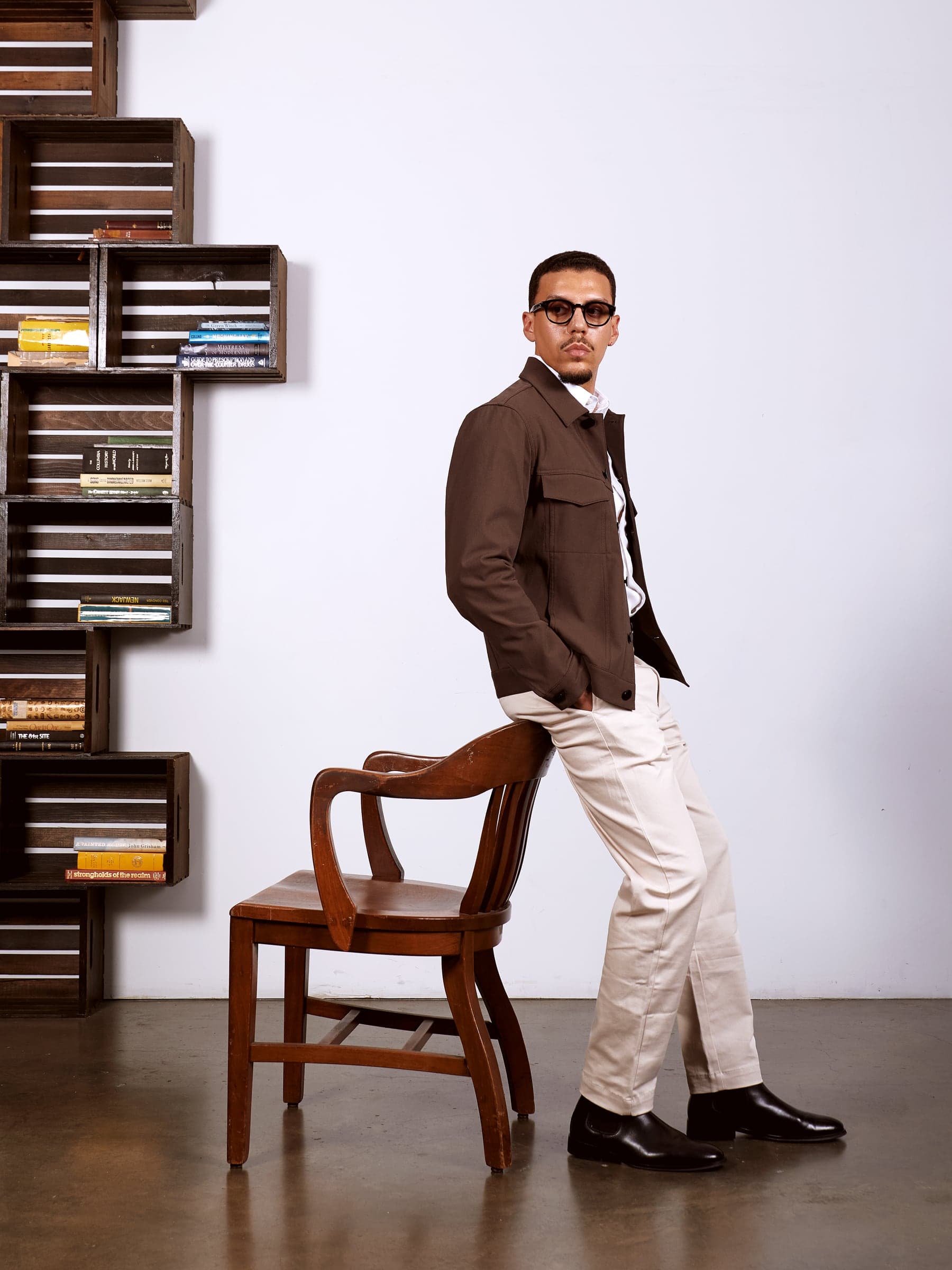 Mars in a brown jacket and cream trousers leaning on a chair beside a wall of books
