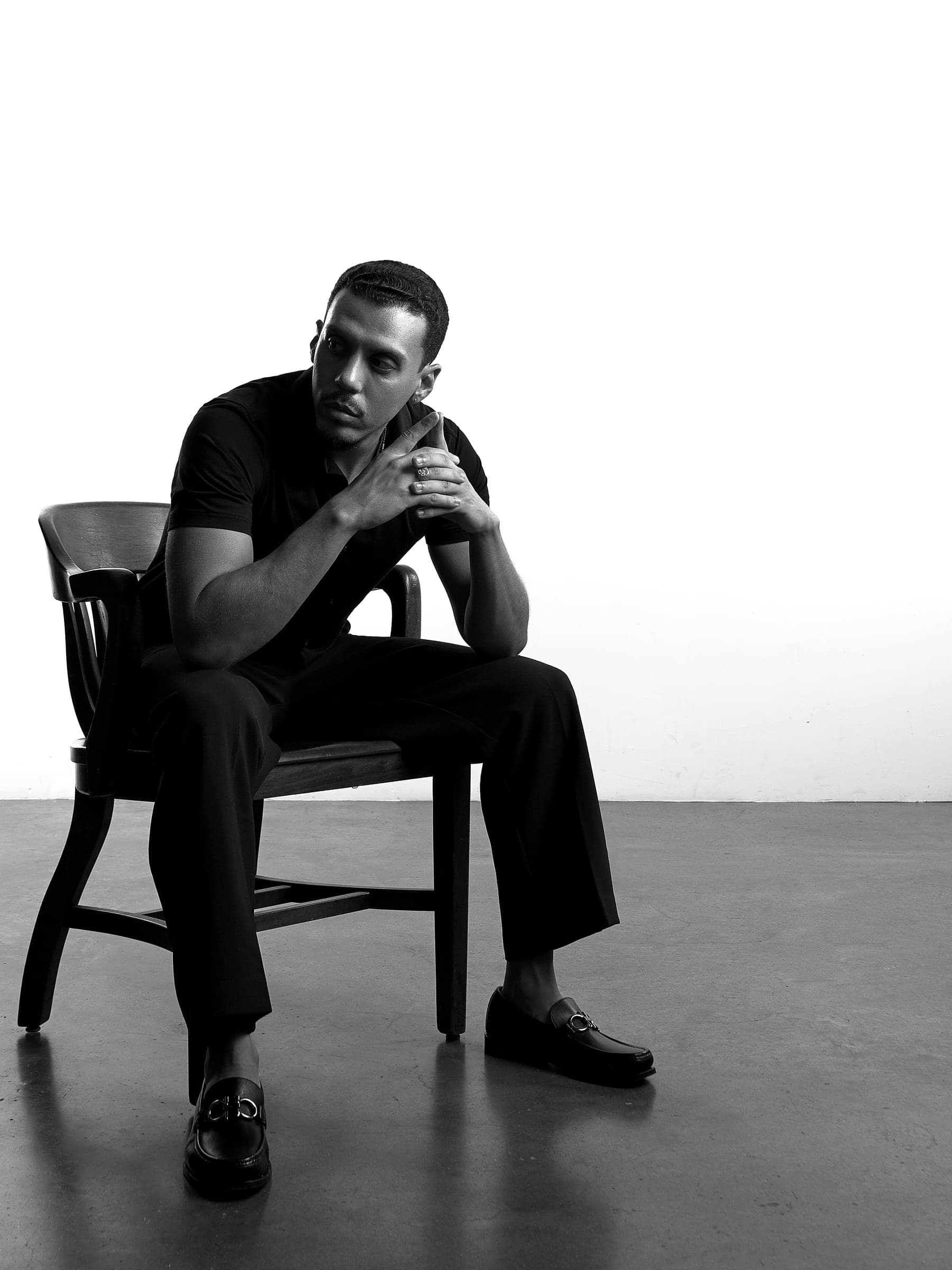 Mars in black and white, seated in a wooden chair, hands clasped, looking down in thought