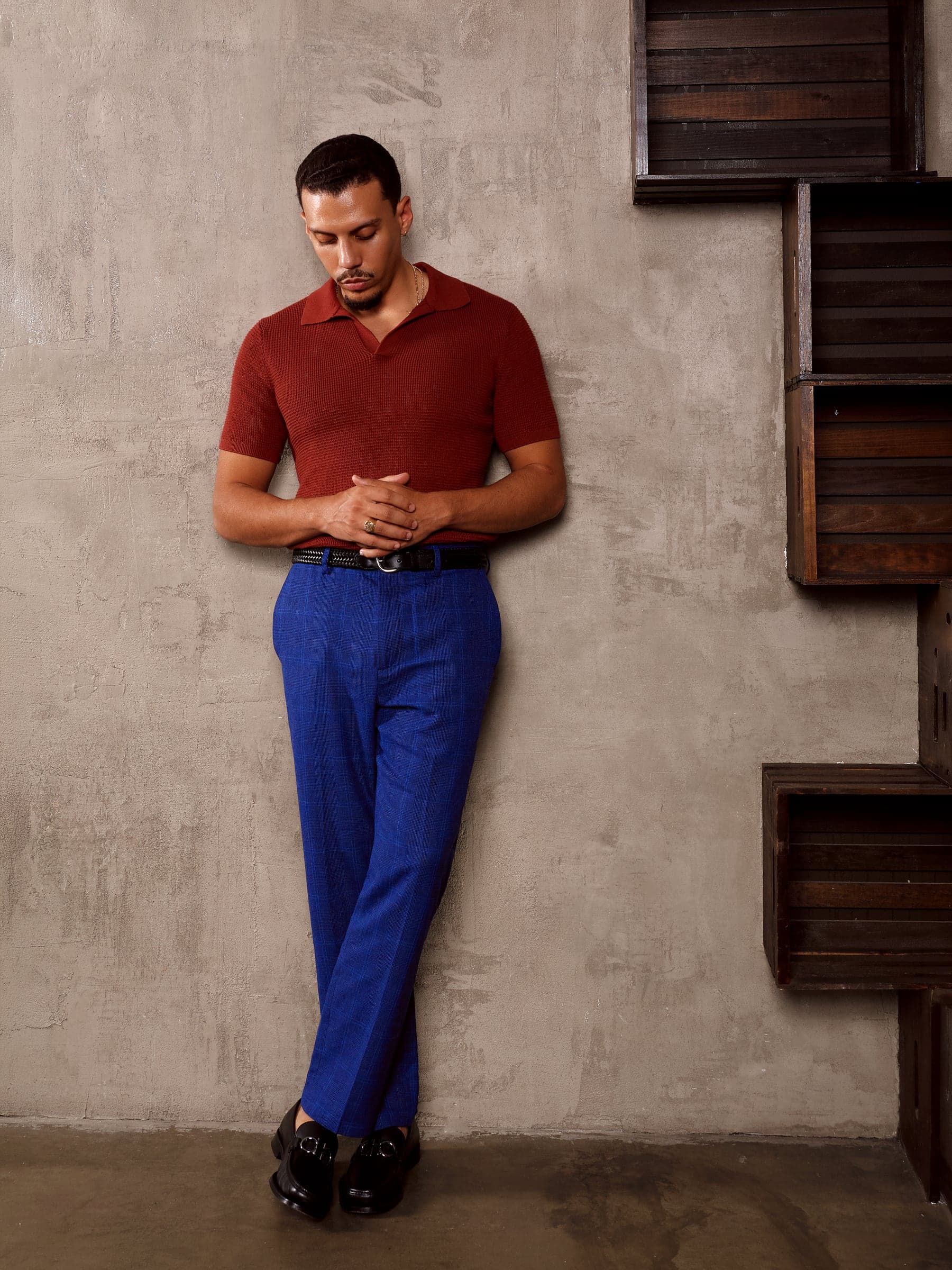 Mars in a red polo shirt and blue trousers standing against a plaster wall beside a wooden shelf